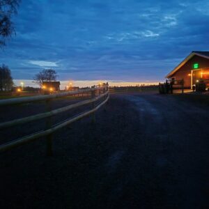 Häststaket vid grusväg och stallbyggnad i kvällsljus